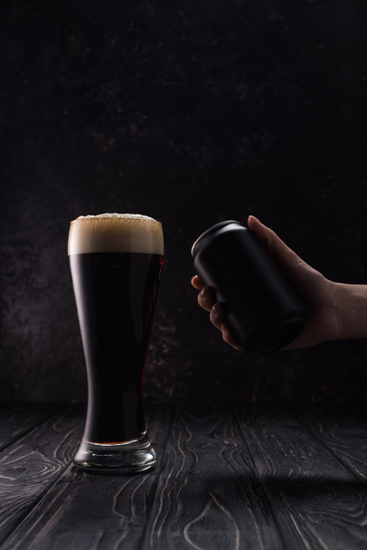 cropped view of man holding black can near glass of dark beer with foam on wooden table - Photo, Image