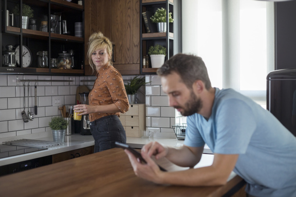 Couple in kitchen. Man is browsing on tablet while woman brings refreshments. - Photo, Image