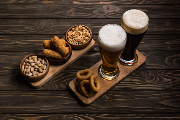 木製のテーブルの上にピーナッツ、ピスタチオ、フライドチーズ、オニオンリングとボウルの近くに暗くて軽いビールのグラス - 写真・画像
