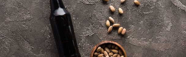 panoramic shot of bottle of dark beer near pistachios on brown textured surface - Foto, Imagen