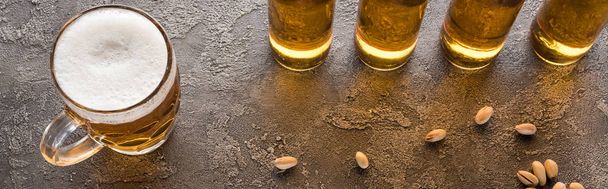 panoramische foto van flessen licht bier in de buurt van verspreide pistachenoten op bruin getextureerde oppervlak - Foto, afbeelding