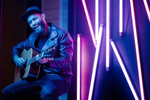 Singer and guitar player singing on a stage with neon lights. He is a rocker and he is wearing leather biker jacket or asymmetric zip jacket with black cap, jeans. - Fotó, kép