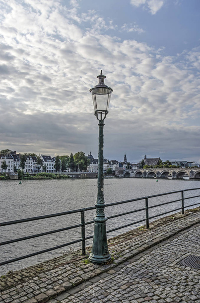 Traditionele lantaarn op Stenenwal (stenen muur) kade op de oostelijke oever van de Maas in Maastricht, met het stadscentrum aan de andere bank op de achtergrond - Foto, afbeelding
