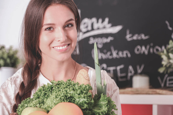 Smiling young woman holding vegetables standing in kitchen. Smiling young woman - Фото, изображение
