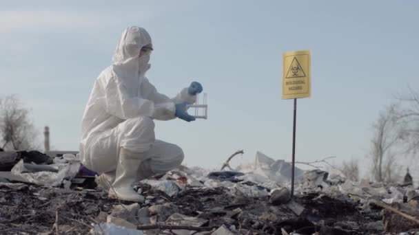 vrouw wetenschapper in uniforme en beschermende handschoenen houden reageerbuisjes nemen monster van afval van de grote vuile dump in de buurt van een teken biologisch gevaar - Video