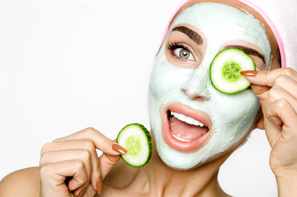 Young smiling woman with a clay mask. Photo of attractive young woman covering her eyes with cucumbers on a white background. Grooming himself - Image - Photo, Image
