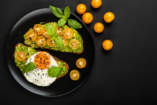 Open sandwiches with avocado guacamole, yellow cherry tomatoes, fried egg and basil on a black plate on the black background. Top view. Copy space. - Photo, Image