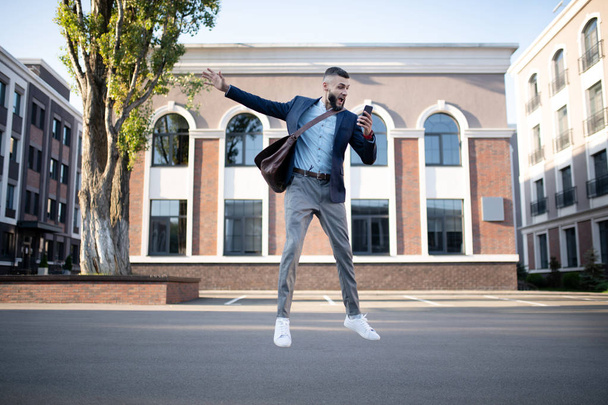 Businessman holding phone jumping while feeling emotional - Фото, изображение