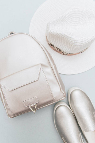 Summer layout of clothes of a young girl. Fashionable silver slip-ons, a leather backpack and a white hat on a pastel blue background - Photo, Image