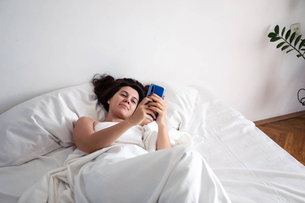 жінка зі смартфоном у ліжку з білими простирадлами
 - Фото, зображення