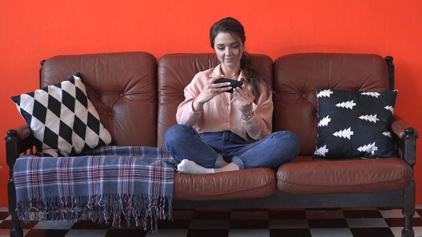 Vista de cerca de la joven mujer bonita en camisa rosa y jeans sentados en el sofá marrón con almohadas y jugar juego móvil por teléfono. Imágenes de archivo. Sensación de felicidad y relax en casa
 - Foto, imagen