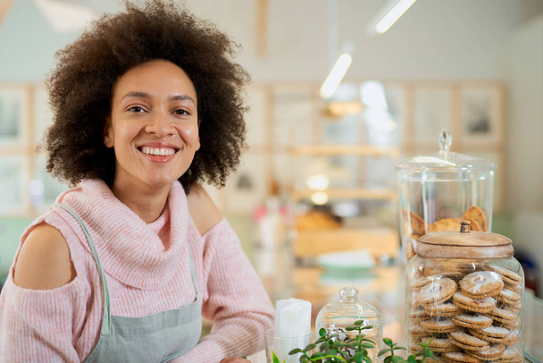 Attractive mixed race employee in pink turtleneck sweater and with apron posing next to jar with cookies. Pastry shop interior. - Foto, Bild