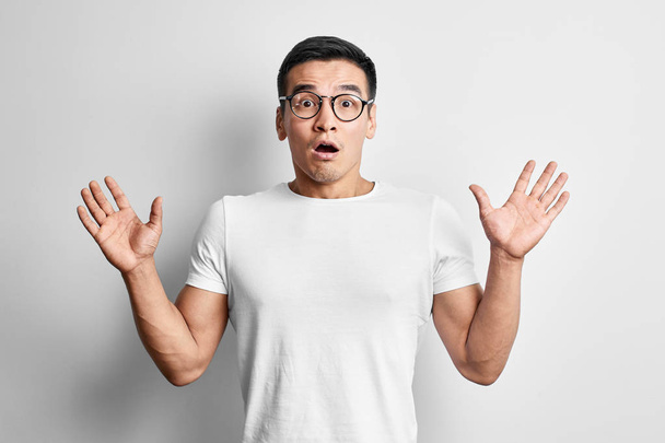 Shocked Asian man dressed in casual and glasses with open mouth and hands up looks to the camera . Surprised young Kazakh student guy on white studio background - Photo, Image