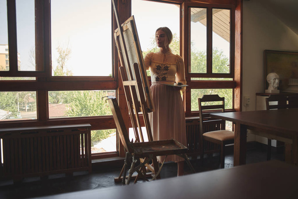 Beautiful talented young woman artist paints a picture with brush and oil on an easel with palette in hand in stylish creative art studio - Foto, imagen