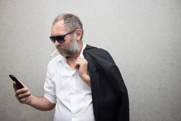 sakallı ve ceketli gözlüklü adam bir telefon ve sigara tutar - Fotoğraf, Görsel