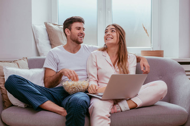 Young guy and woman eating popcorn and watching movie from notebook in a cozy living room - Photo, Image