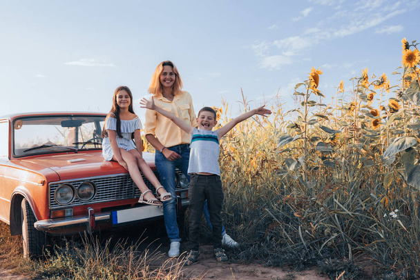 Famille heureuse près de champ de tournesols, maman avec deux enfants, jeune fille en robe assise sur le capot de la voiture rétro rouge, mère et fils debout à côté, vacances d'été
 - Photo, image