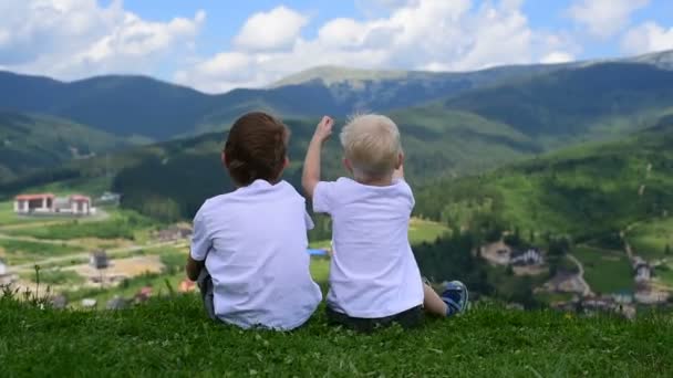 Twee kleine jongens zitten en spelen op een groene weide tegen de achtergrond van een dorp en bergen. - Video
