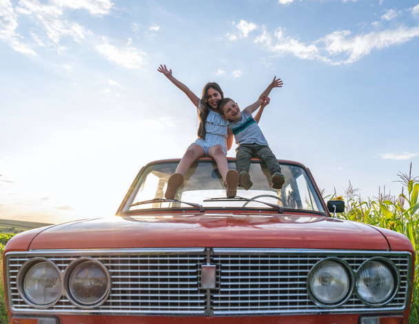 Happy smiling teen children in casual clothes sitting together on the roof of the red retro car huging and holding arms raised, blue sky and sunset on the background - Foto, imagen