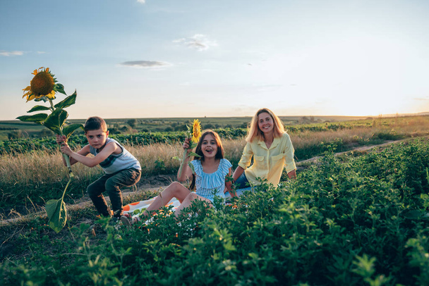 Happy funny smiling family on the picnic, serious boy in casual clothes crouched with sunflower in hands, her mom in yellow shirt with teen sister in blue striped dress are sitting on plaid among the - Photo, Image