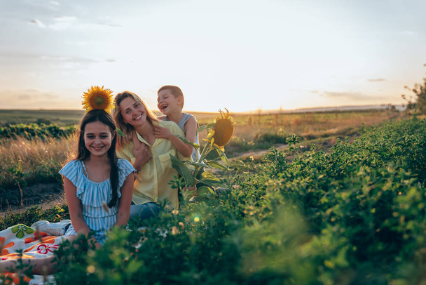 Happy figlarny rodzina siedzi na kratę wśród posiłków, brunetka Teen Girl w niebieskich pasiastej sukni ze słonecznikiem na głowie i jej mama w żółtej koszulce z małym bratem korzystających piknik razem - Zdjęcie, obraz