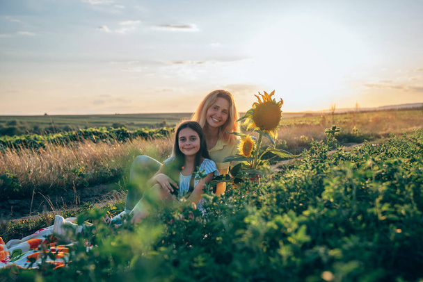 Szczęśliwy uśmiechnięty młody blond mama w żółtej koszulce siedzi na kratę wśród posiłków z jej Nastoletnia Córka brunetka w paski niebieska sukienka, zachód słońca na tle - Zdjęcie, obraz