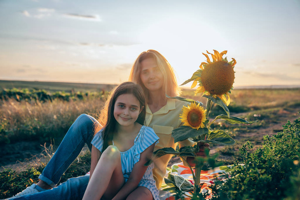 Hermosa chica adolescente en vestido azul sentado en el cuadros entre el mealow cerca de girasoles con su joven madre rubia en camisa amarilla, puesta de sol en el fondo
 - Foto, Imagen