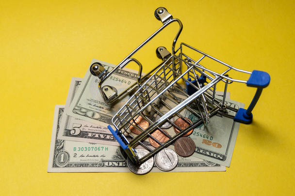 An inverted metal shopping trolley lies on small US bills and coins. The concept of online purchases and sales, unsuccessful transactions and a price flip. Dollars and cents on a yellow background. - Photo, image