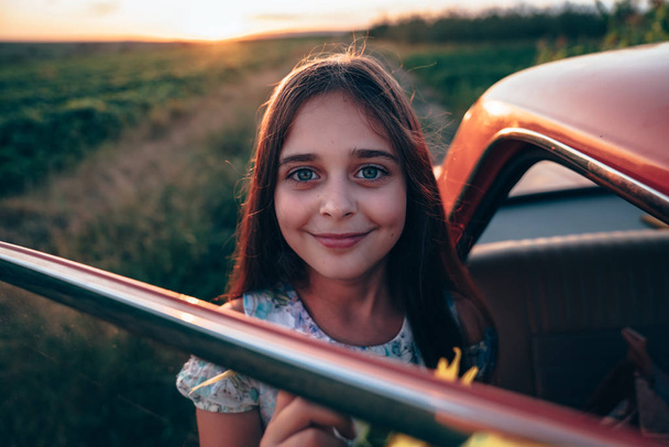 Close up portrait of beautiful brunette teen girl with natural healthy skin and deep blue eyes that standing near the red car and looking at the camera, sunset on the background - Фото, изображение