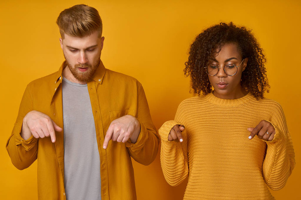 Stupefied young mixed race women and caucasian man point at floor together, have surprised gaze down, dressed casually, stand next to each other against an orange background. Wow, just look there - Photo, image