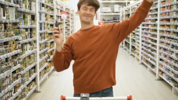 happy guy in brown sweatshirt dances holding smartphone in shop - Footage, Video