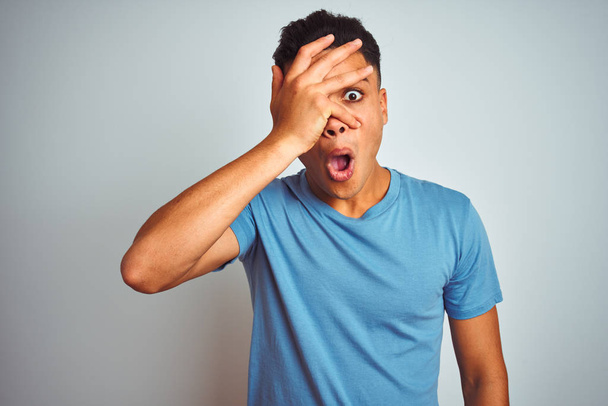 Jonge Braziliaanse man dragen blauw t-shirt staande over geïsoleerde witte achtergrond gluren in shock dekking van gezicht en ogen met de hand, kijkend door vingers met beschaamde uitdrukking. - Foto, afbeelding