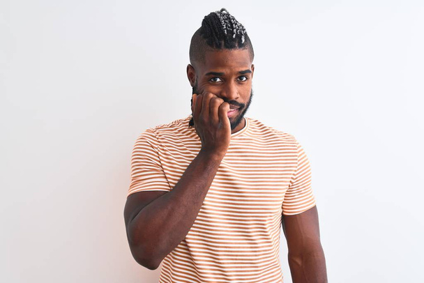 Afro-Amerikaanse man met vlechten draagt gestreept t-shirt over geïsoleerde witte achtergrond kijken gestrest en nerveus met handen op de mond bijtende nagels. Angst probleem. - Foto, afbeelding