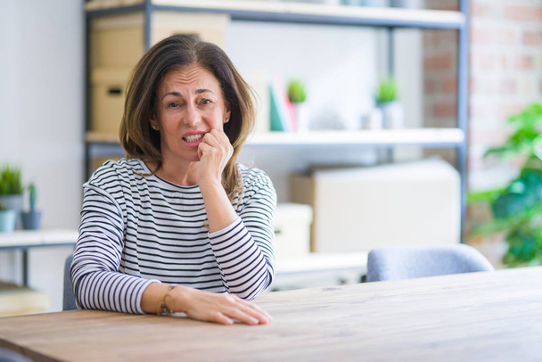 Orta yaş yaşlı kadın evde masada oturan stresli ve sinirli ağız tırnakısırma elleri ile bakıyor. Anksiyete sorunu. - Fotoğraf, Görsel