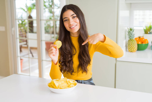 Young woman eating fastfood potato chips with surprise face pointing finger to himself - Photo, Image