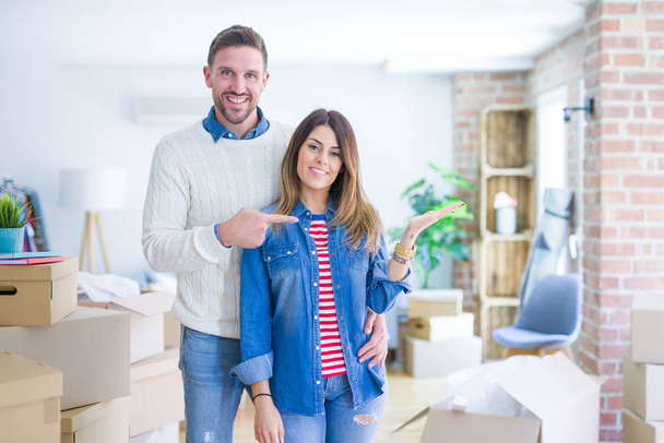 Young beautiful couple standing at new home around cardboard boxes amazed and smiling to the camera while presenting with hand and pointing with finger. - Foto, Imagem