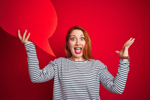 Young beautiful redhead woman holding speech bubble over red isolated background very happy and excited, winner expression celebrating victory screaming with big smile and raised hands - Foto, Imagen