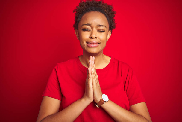 Joven mujer afroamericana hermosa con pelo afro sobre fondo rojo aislado rezando con las manos juntas pidiendo perdón sonriendo confiado
. - Foto, Imagen