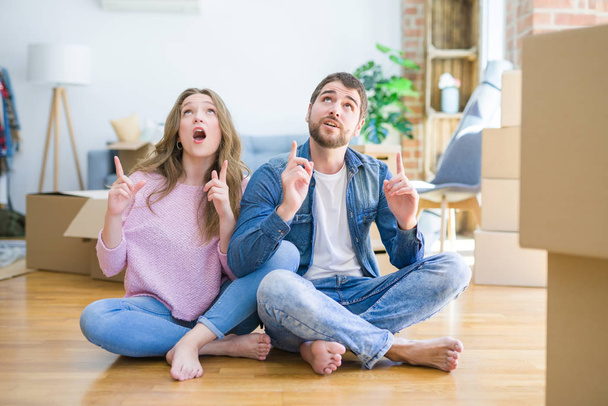 Young beautiful couple moving to a new house sitting on the floor amazed and surprised looking up and pointing with fingers and raised arms. - Photo, Image