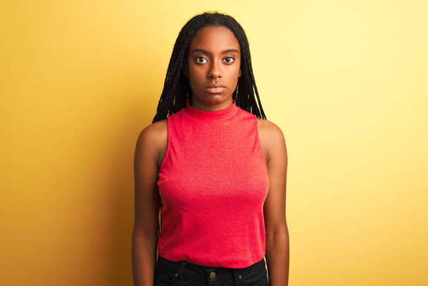Африканская американка в красной футболке, стоящей на изолированном желтом фоне, расслабленная с серьезным выражением лица. Простой и естественный взгляд на камеру
. - Фото, изображение