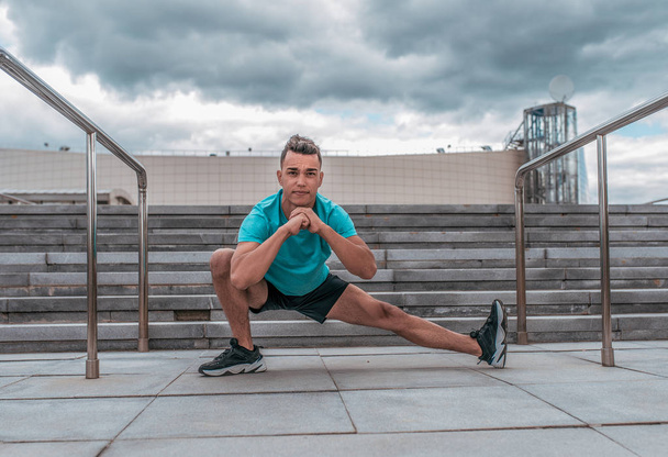Male athlete doing leg warms before fitness workout workout, jogging in summer morning in city. Sportswear Sneakers Shorts. Active lifestyle of youth. - 写真・画像