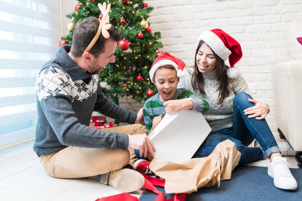 Ευτυχισμένοι γονείς με ενθουσιασμένο γιο που κοιτάζει το χριστουγεννιάτικο δώρο στο καθιστικό - Φωτογραφία, εικόνα