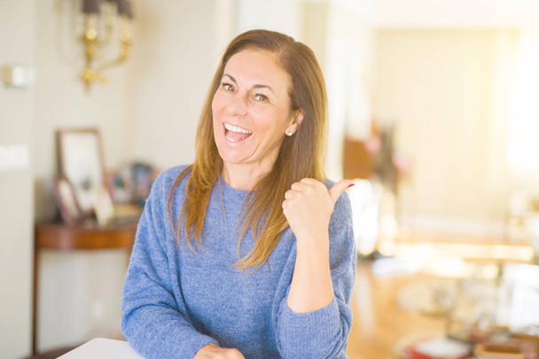 Beautiful middle age woman at home smiling with happy face looking and pointing to the side with thumb up. - 写真・画像