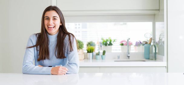 Groothoek foto van mooie jonge vrouw zittend op witte tafel thuis met een gelukkige en koele glimlach op het gezicht. Gelukkige persoon. - Foto, afbeelding