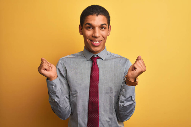 Молодой привлекательный арабский бизнесмен в рубашке и галстуке на изолированном желтом фоне радовался и удивлялся успеху с поднятыми руками и открытыми глазами. Концепция победителя
. - Фото, изображение