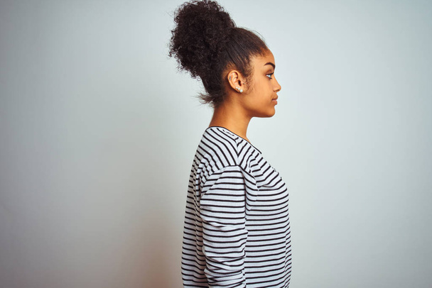 Африканская американка в полосатой футболке, стоящей на изолированном белом фоне, глядя в сторону, расслабиться профиль позировать с естественным лицом и уверенной улыбкой
. - Фото, изображение
