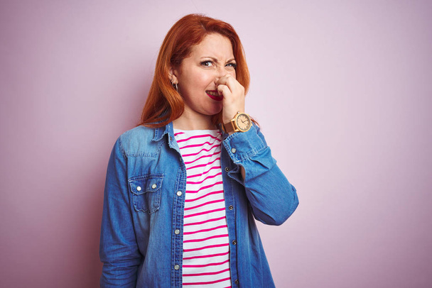 Beautiful redhead woman wearing denim shirt and striped t-shirt over isolated pink background smelling something stinky and disgusting, intolerable smell, holding breath with fingers on nose. Bad smells concept. - Fotoğraf, Görsel