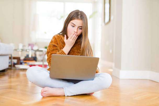 Beautiful young girl studying using laptop sitting on the floor at home cover mouth with hand shocked with shame for mistake, expression of fear, scared in silence, secret concept - Φωτογραφία, εικόνα