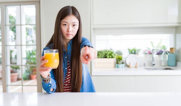 Gyönyörű ázsiai nő iszik egy pohár friss narancslevet ujjal mutogatva a kamerára és rád, kézjel, pozitív és magabiztos gesztus elölről - Fotó, kép
