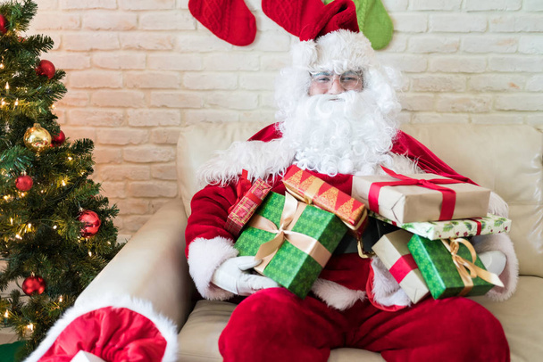 Санта-Клаус сидит с подарочными коробками на диване в украшенном доме во время Рождества
 - Фото, изображение